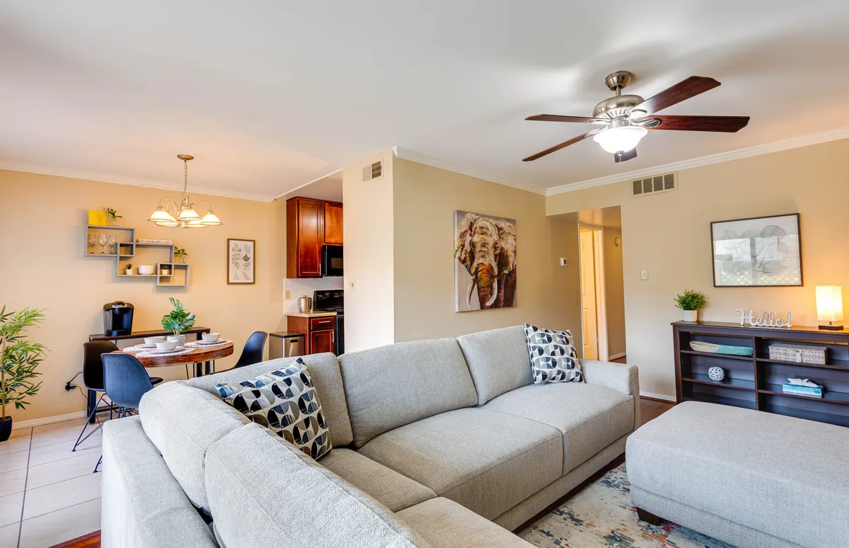 An open, inviting living space showcasing a plush sofa, dining table, artistic decor on the walls, and a visible kitchen area in the background. The space is bright and seamlessly integrates lounging, dining, and cooking zones, with cozy furnishings and modern touches.