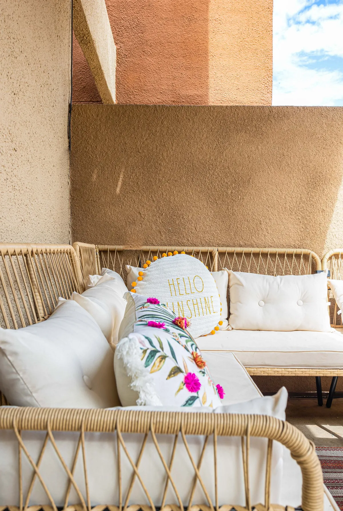 A cheerful outdoor patio scene featuring woven furniture with decorative pillows set against a textured wall. The area is bathed in sunlight, creating an inviting atmosphere for relaxation and socializing. The open and spacious layout makes this patio an ideal spot for both dog owners and their pets to relax safely outdoors.