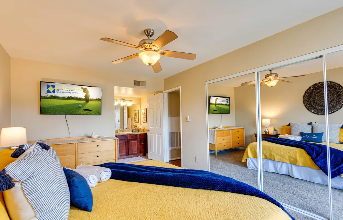 A stylish bedroom with a yellow and blue bedspread, a television displaying a golf scene, and a large mirror reflecting the room’s brightness. The combination of vibrant bedding and soft lighting creates a welcoming, restful atmosphere.
