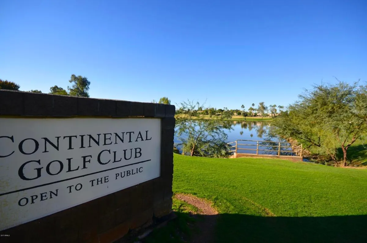 A serene golf course scene featuring a golf club sign, calm lake, lush green grass, and palm trees in the background. The area is inviting, tranquil, and perfectly maintained, with ample open space for golfers and nature lovers alike.