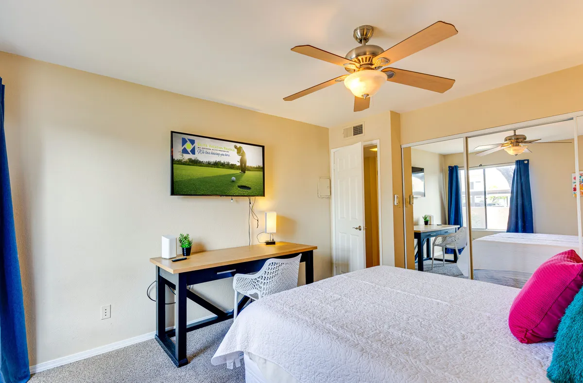 A serene bedroom featuring a plush bed with crisp white bedding, a dedicated desk and comfortable chair, and a flat-screen TV mounted on the wall. Ample natural light filters through the window, creating a peaceful and functional space ideal for both rest and productivity.