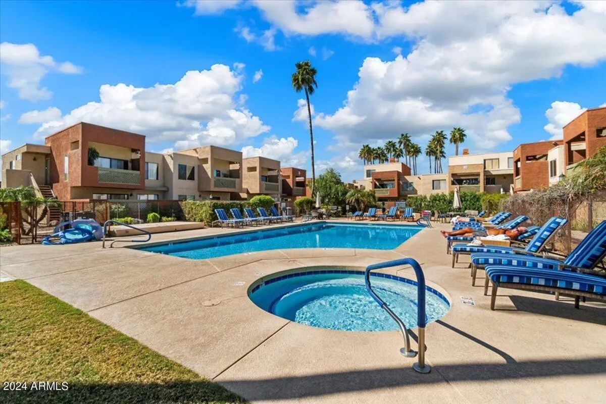 A sparkling swimming pool is the centerpiece of this lively outdoor space, complemented by a bubbling hot tub, inviting lounge chairs, and mature palm trees swaying around the pool deck. Surrounding buildings and lush greenery frame the area, evoking an inviting resort-like atmosphere.