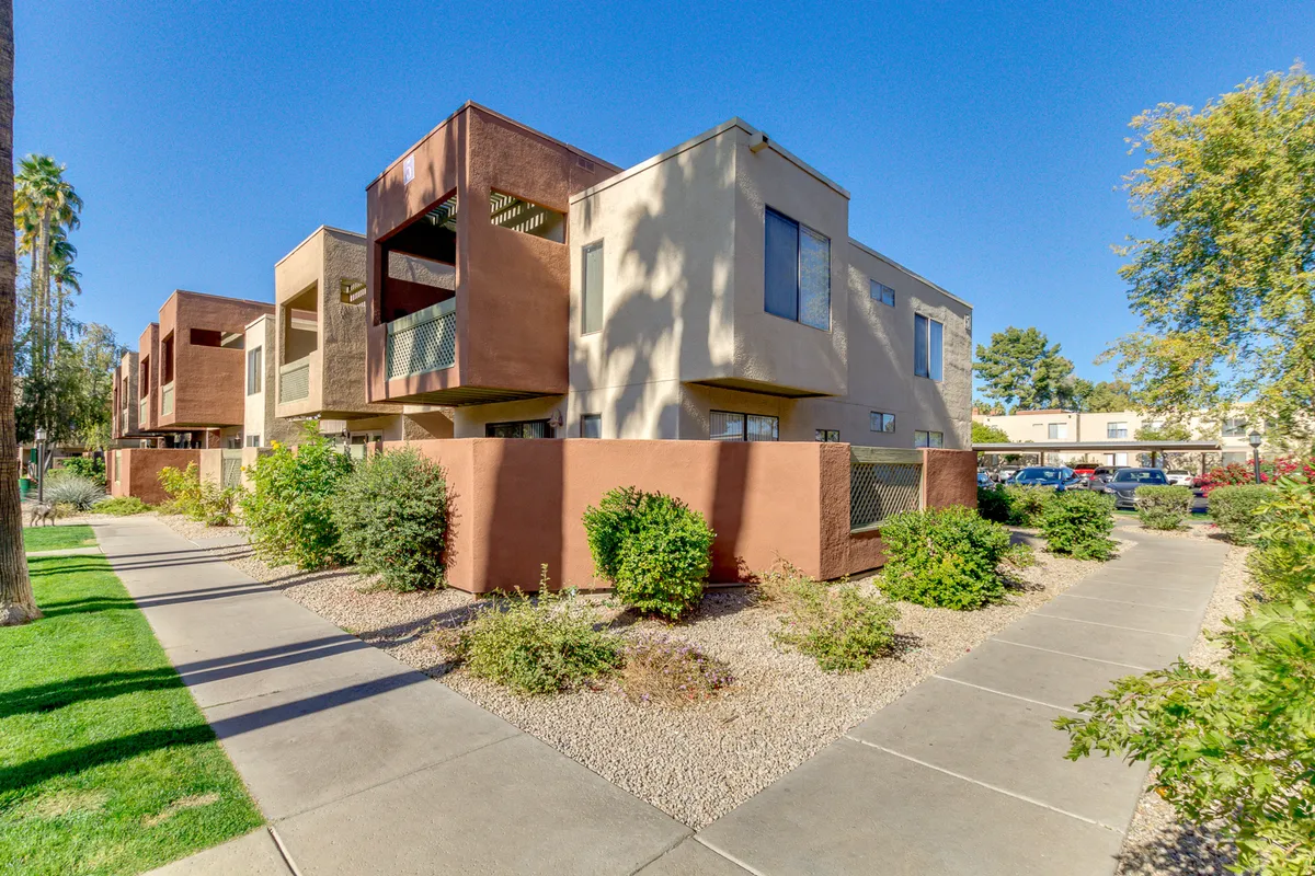 Landscaped pathways bordered by green shrubbery and tall palm trees lead through a modern apartment community. The scene radiates tranquility, showing residents walking and socializing in sunlit outdoor spaces.