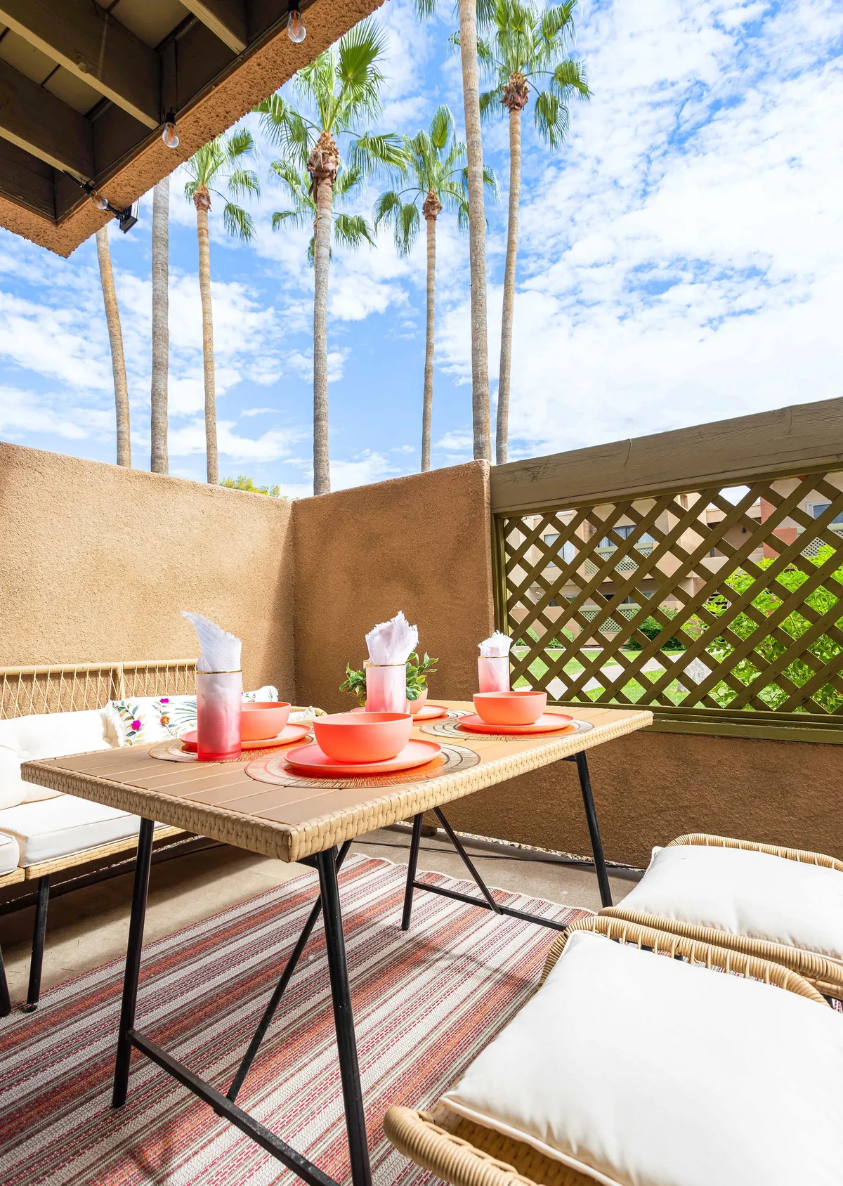 The scene includes a patio dining table set with bright orange dishes, surrounded by a comfortable seating area and the calming presence of palm trees in the background. The space exudes warmth and relaxation, ideal for socializing or unwinding in the open air.