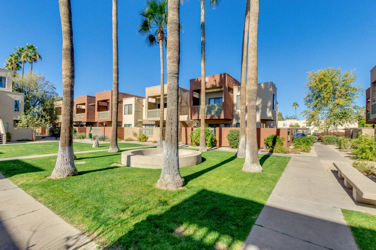 A wide, well-maintained pathway winds through vibrant green grass and under the shade of tall palm trees, with modern apartment buildings nearby. The inviting walkway encourages strolls, socializing, and enjoying the community's lush, open green spaces.